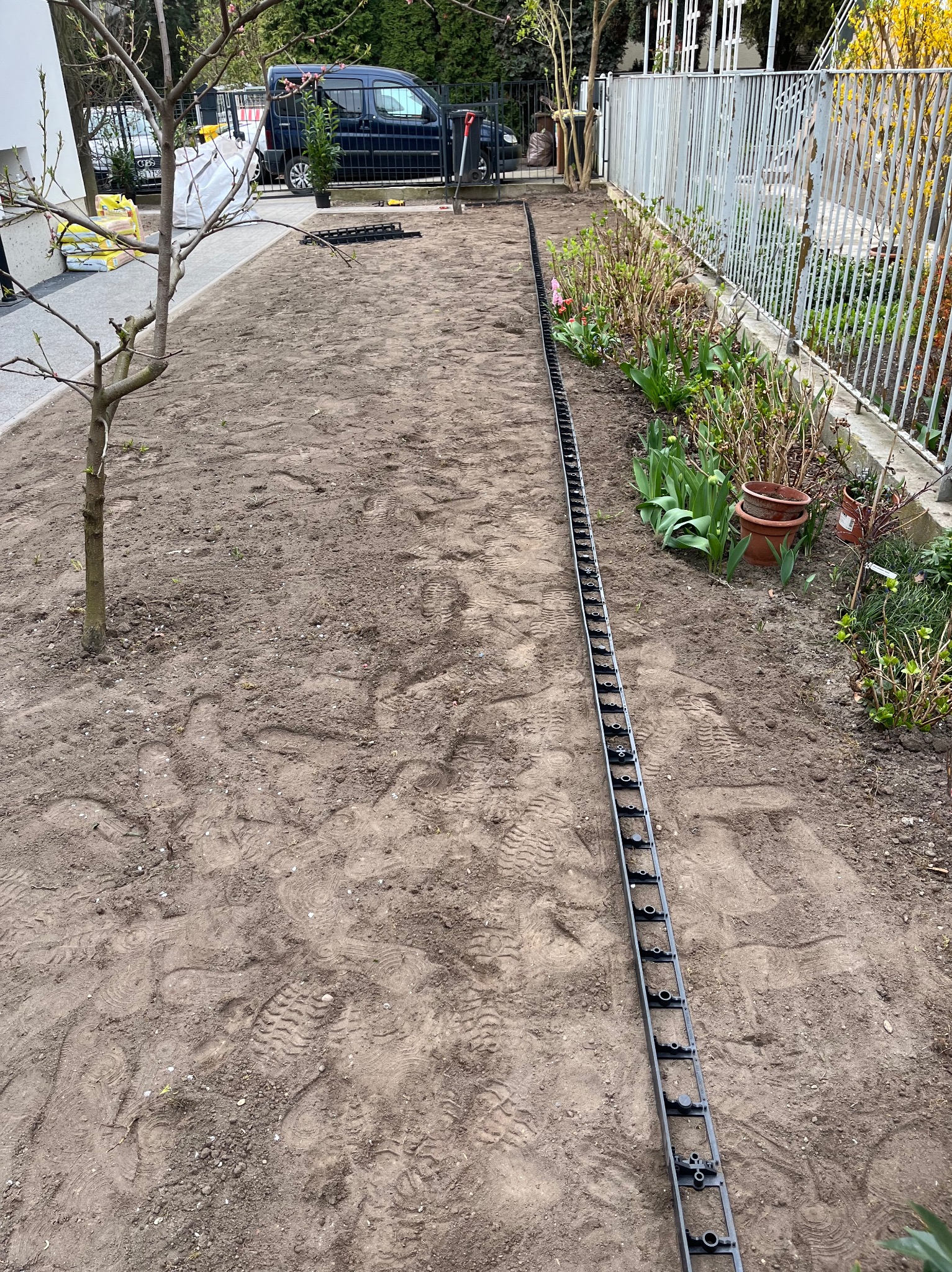 Przygotowany teren pod trawnik z widocznymi elementami systemu drenażowego wzdłuż ogrodzenia, świeżo posadzonymi roślinami i workami z ziemią w tle.