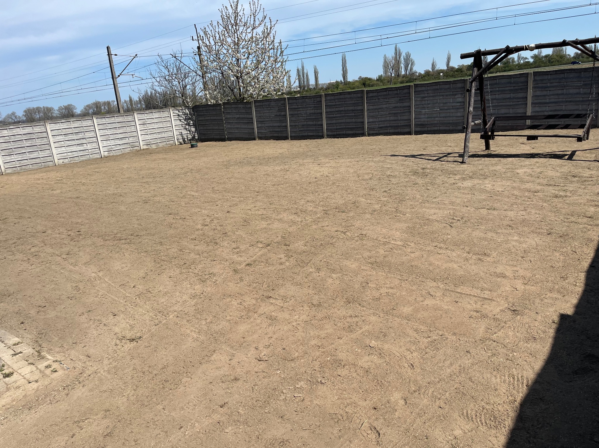 Przygotowany teren pod przyszły ogród, ogrodzenie z betonowych płyt, drewniana huśtawka w prawym rogu, kwitnące drzewo i słupy trakcji elektrycznej w tle.