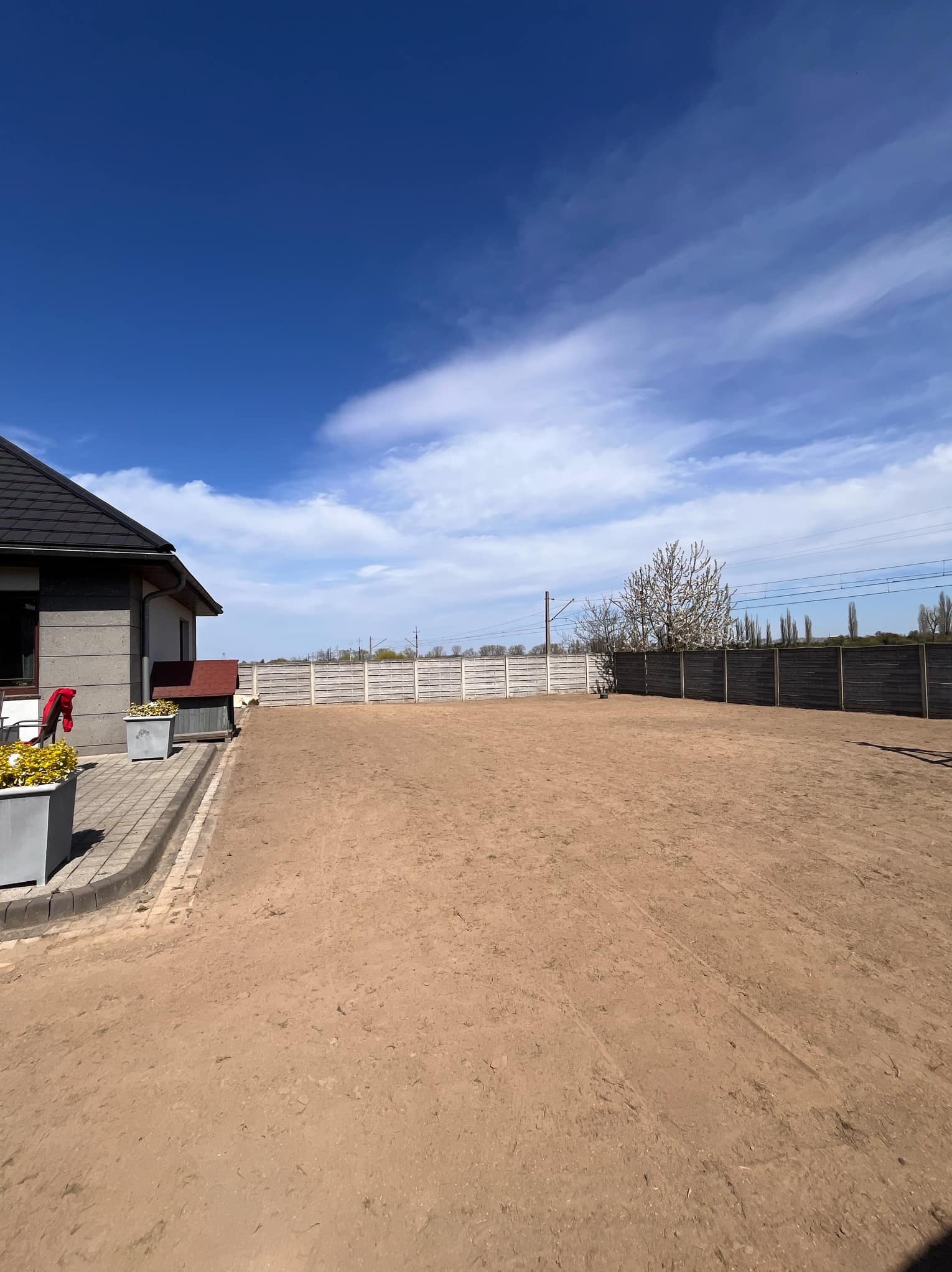 Przygotowany teren pod trawnik obok domu jednorodzinnego, widoczne ślady po bronowaniu, w tle ogrodzenie i linia kolejowa.