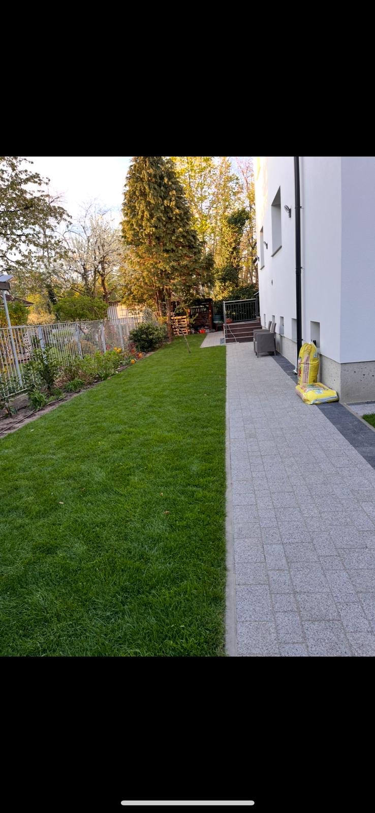 Świeżo założony trawnik z rolki przylegający do jasnej kostki brukowej przy budynku. Widoczne worki z ziemią, sugerujące prace wykończeniowe.