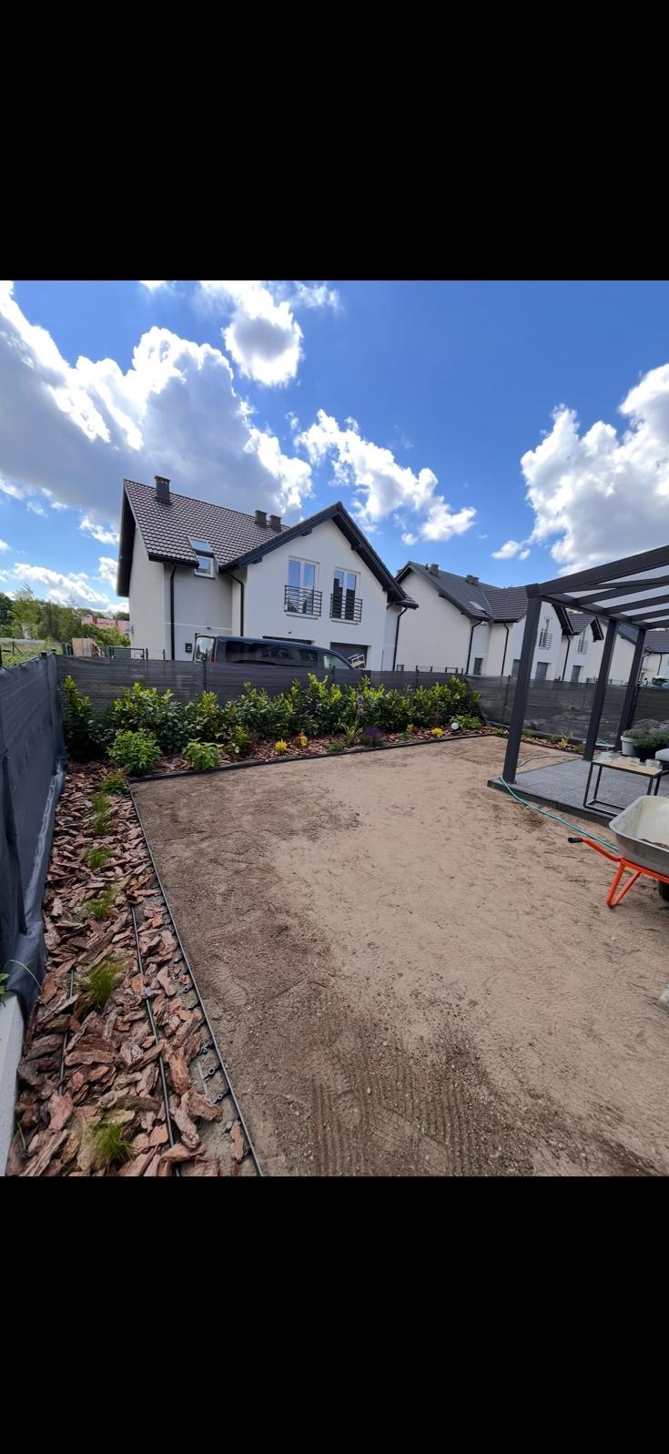 Przygotowany teren pod trawnik z rolki, obrzeża z kory, widoczne nasadzenia krzewów i drzewek, pergola w tle, dom jednorodzinny. Błękitne niebo z chmurami.