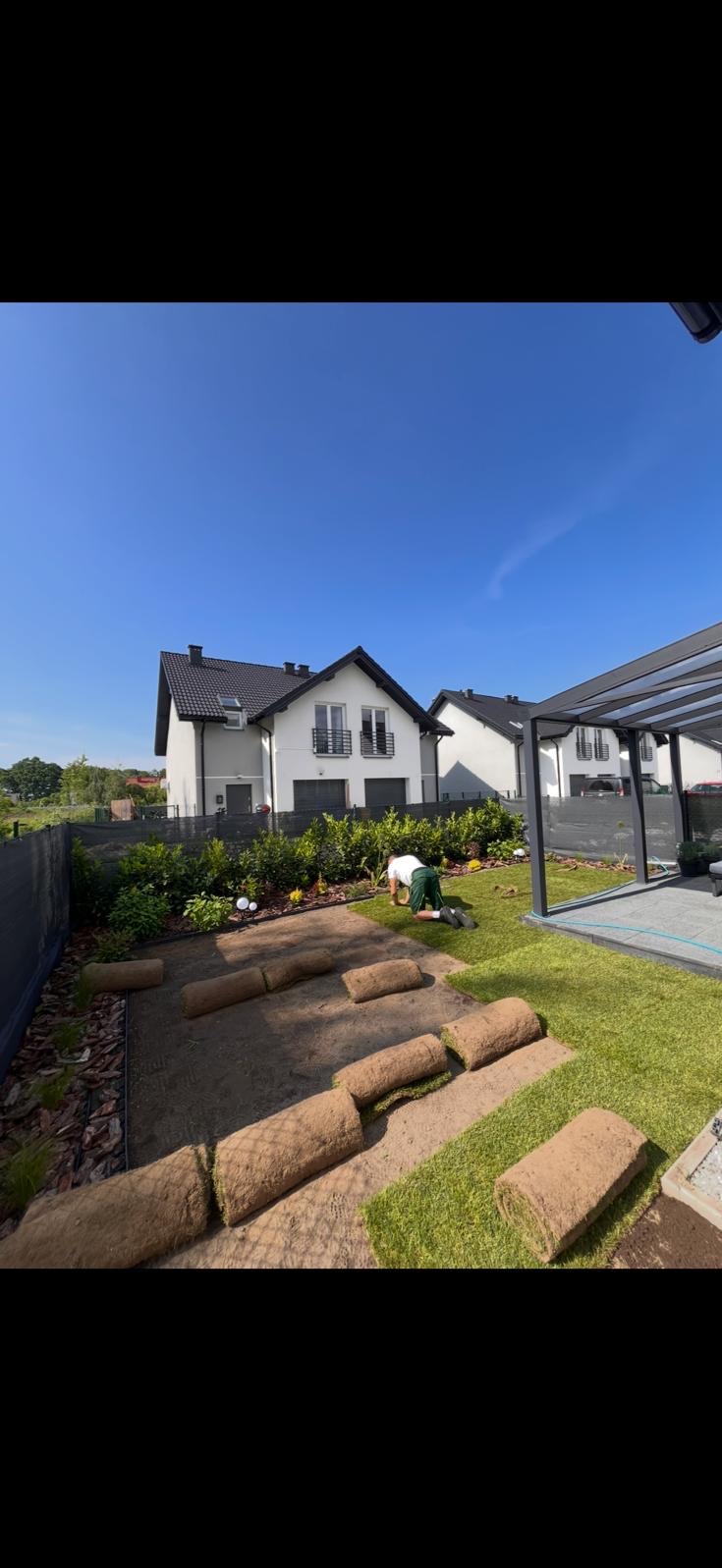 Układanie trawy z rolki w ogrodzie przydomowym. Widoczne rozłożone rolki trawy, pracownik w trakcie pracy oraz fragment już ułożonego trawnika. W tle dom i pergola.