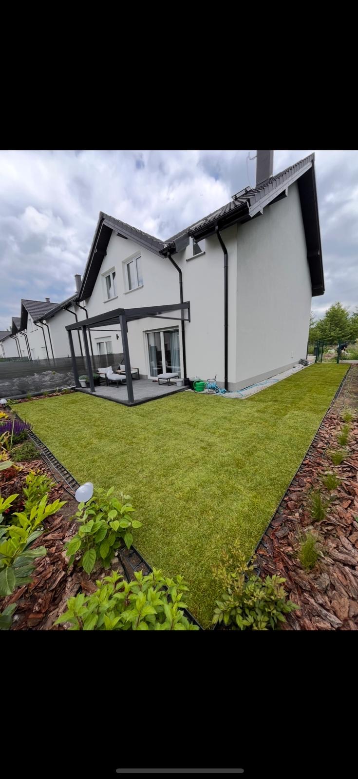 Nowoczesny ogród z równo ułożoną trawą, obrzeżami z kory i elegancką pergolą przy białym domu. Widoczne detale wykończenia i zadbana zieleń.