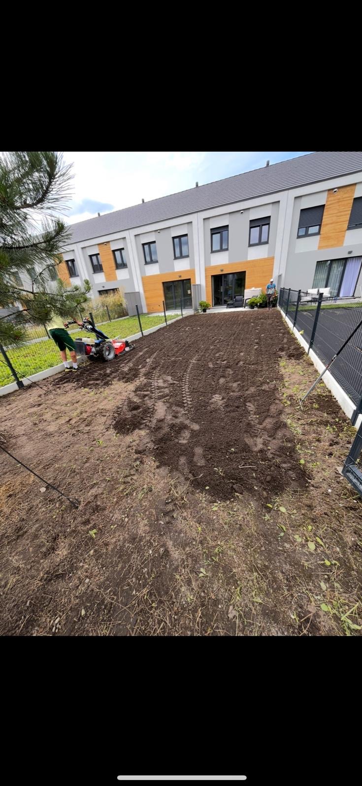 Przygotowanie gruntu pod trawnik: gleba spulchniona glebogryzarką, widoczne ślady opon, w tle budynek mieszkalny z elementami drewnianymi.