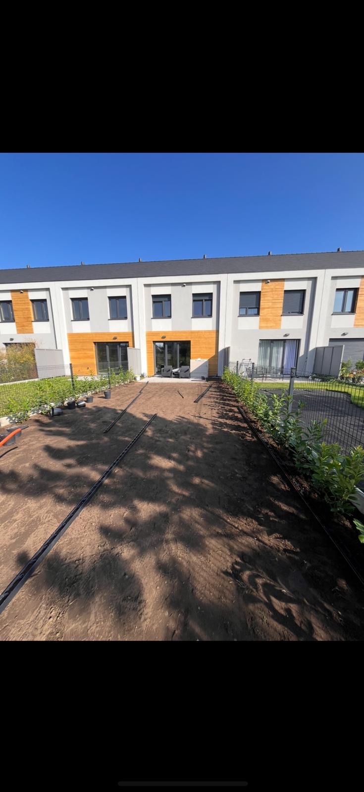 Przygotowany teren pod trawnik z widocznymi liniami nawadniania, obok posadzone niskie krzewy, w tle budynek mieszkalny z drewnianymi elementami elewacji.