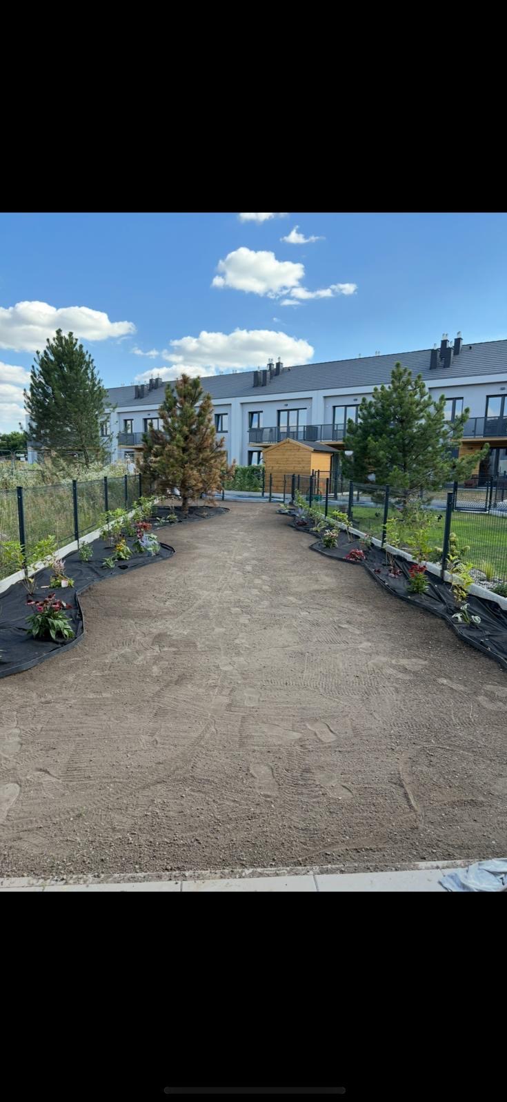 Przygotowany teren pod trawnik z rolki, obrzeża wyłożone agrowłókniną i posadzone kwiaty, w tle budynki mieszkalne i drzewa iglaste.