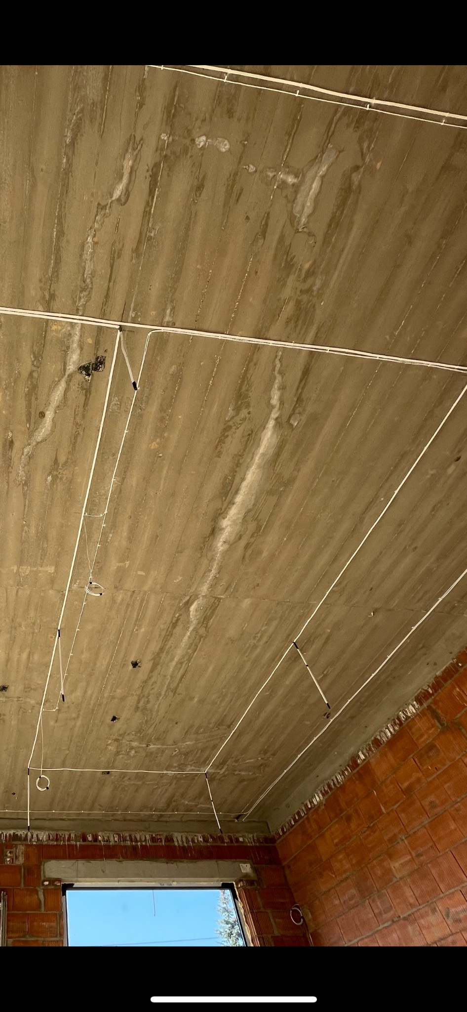 Surowy betonowy sufit z widocznymi przewodami elektrycznymi przymocowanymi do niego, w tle fragment ściany z czerwonej cegły i okno z widokiem na niebo.