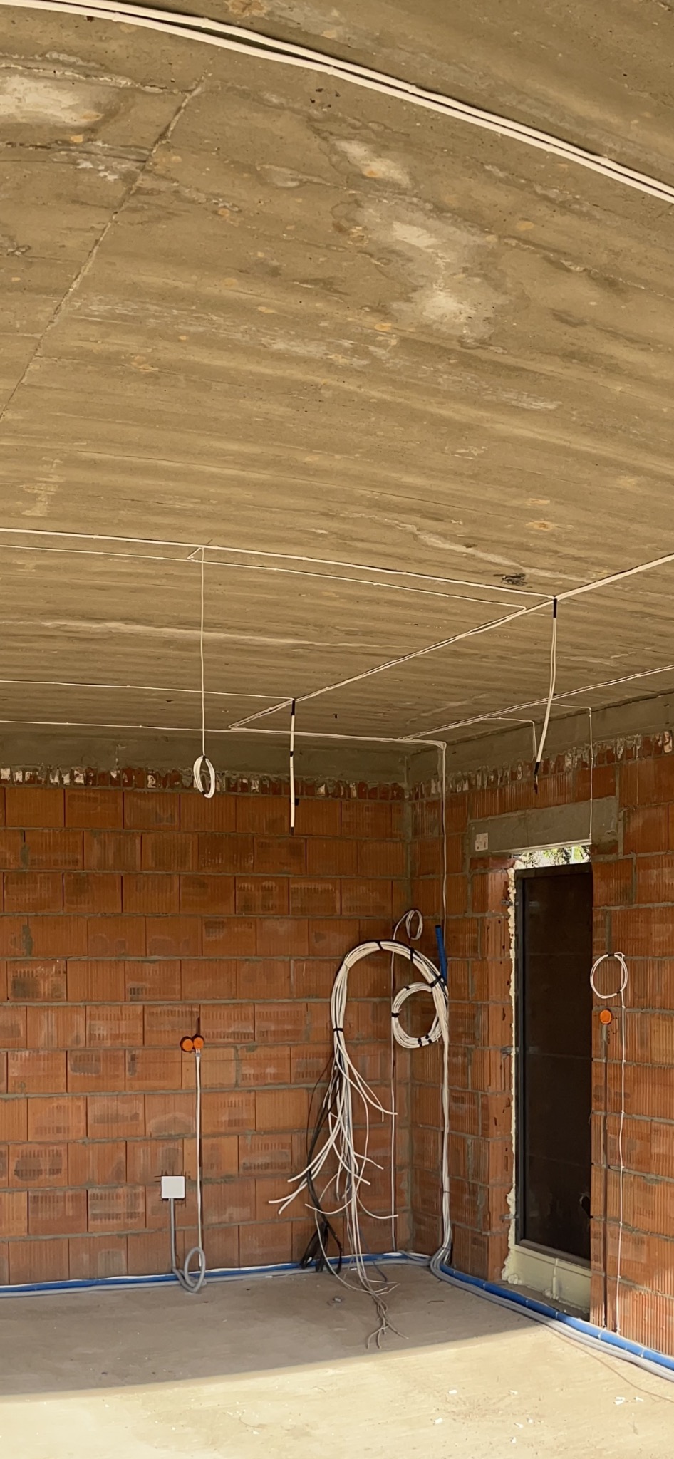 Surowy stan wnętrza domu w trakcie prac. Widoczne przewody elektryczne prowadzone po suficie i ścianach z cegły, puszki instalacyjne oraz zwisające kable. Nieotynkowany betonowy sufit.