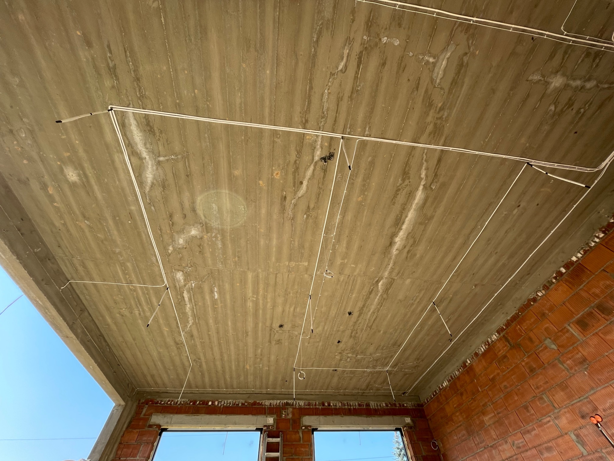 Surowy betonowy sufit z widocznymi, częściowo zamontowanymi przewodami elektrycznymi w nowo budowanym budynku z ceglanymi ścianami i oknami.