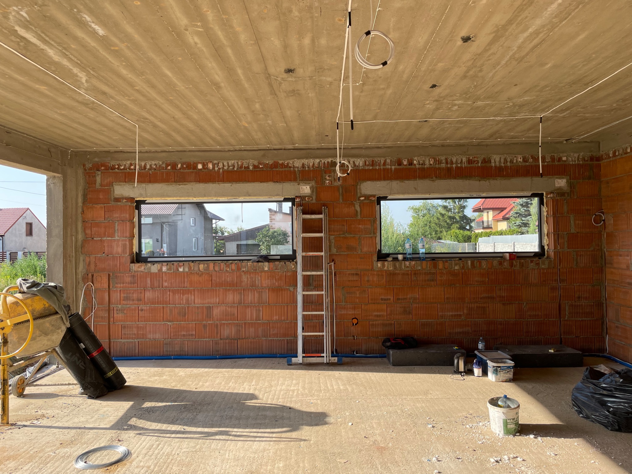 Wnętrze surowego budynku w trakcie instalacji elektrycznej: widoczne przewody pod sufitem, ceglane ściany z oknami, drabina, mieszalnik do betonu i materiały budowlane.