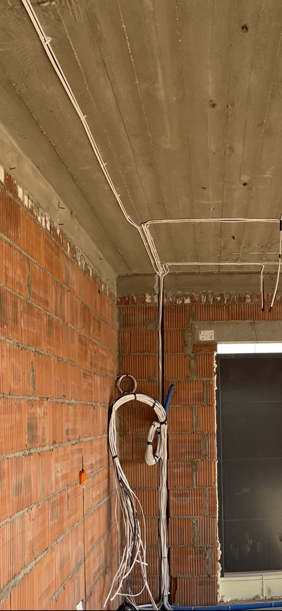 Surowe wnętrze w trakcie prac: widoczne przewody elektryczne prowadzone po suficie i ścianach z czerwonej cegły, tymczasowo przymocowane do ściany, z niebieską rurką karbowaną w tle.