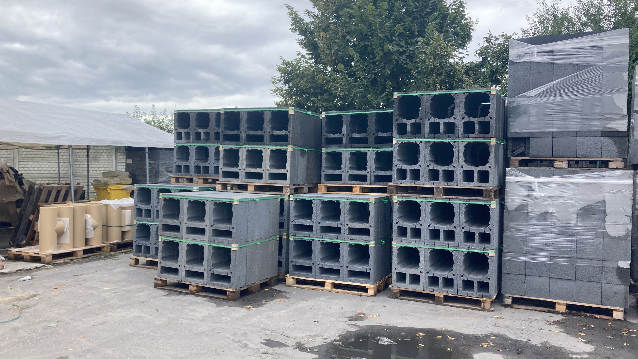 Szare prefabrykaty betonowe z otworami ułożone na drewnianych paletach, część zafoliowana, na zewnątrz, w tle drzewa i biały namiot.