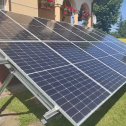 Panele słoneczne zamontowane na metalowej konstrukcji przed domem z czerwonej cegły i beżową elewacją, widoczne skrzynki z czerwonymi kwiatami pod oknami.