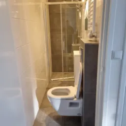 Nowoczesna łazienka z wiszącą toaletą, kabiną prysznicową ze szklanymi drzwiami i grzejnikiem drabinkowym, wykończona w odcieniach bieli i szarości.