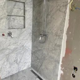 Prysznic wyłożony marmurowymi płytkami z wnęką na kosmetyki i odpływem liniowym, widoczne niedokończone prace elektryczne po prawej stronie.