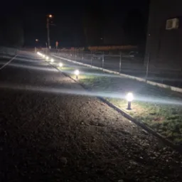 Rząd kulistych lamp oświetlających żwirową drogę wzdłuż ogrodzenia posesji w nocnym krajobrazie.
