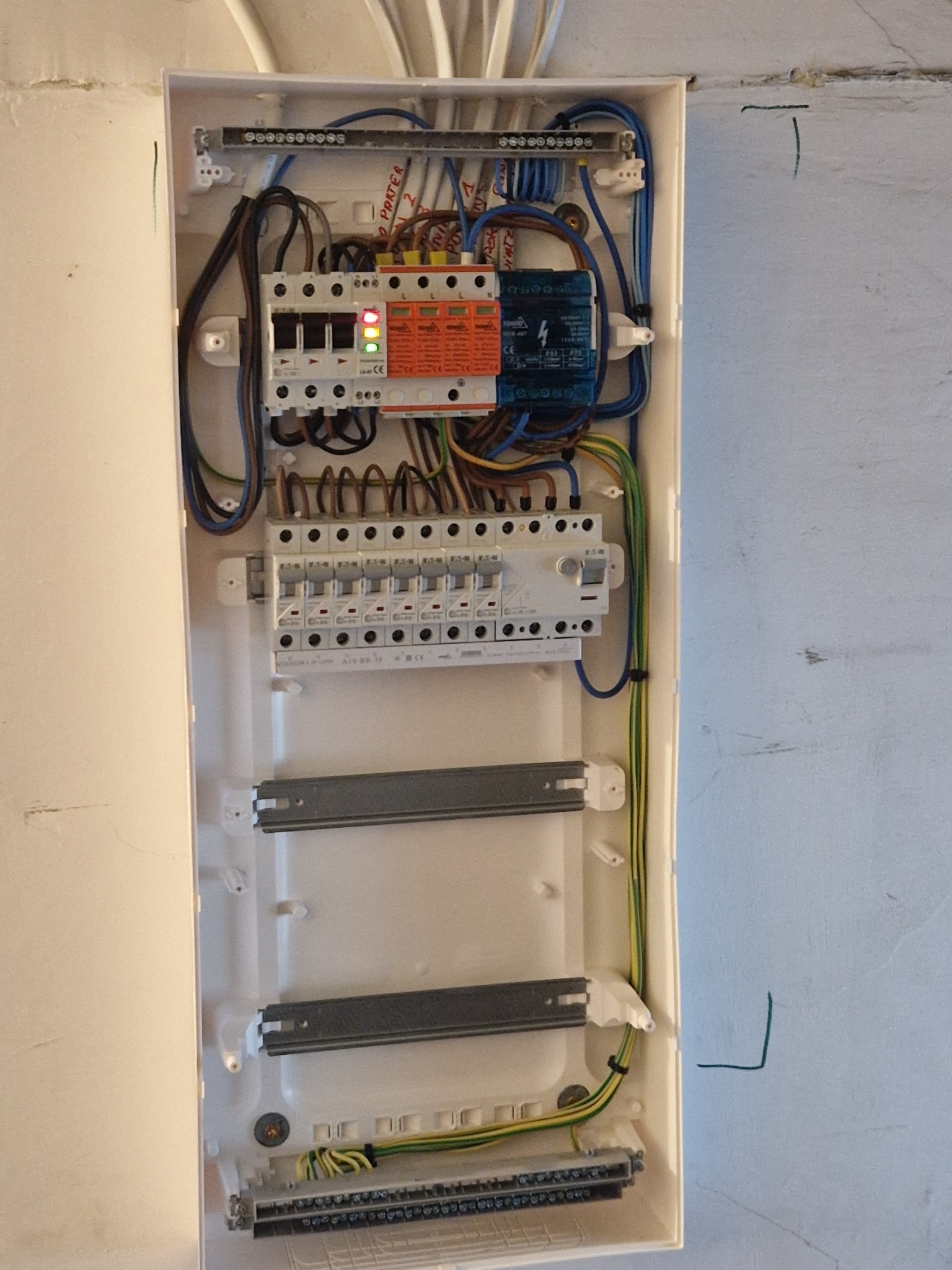 Otwarta skrzynka rozdzielcza z widocznymi bezpiecznikami, przewodami i listwami zaciskowymi, zamontowana na ścianie z widocznym śladem po ołówku.