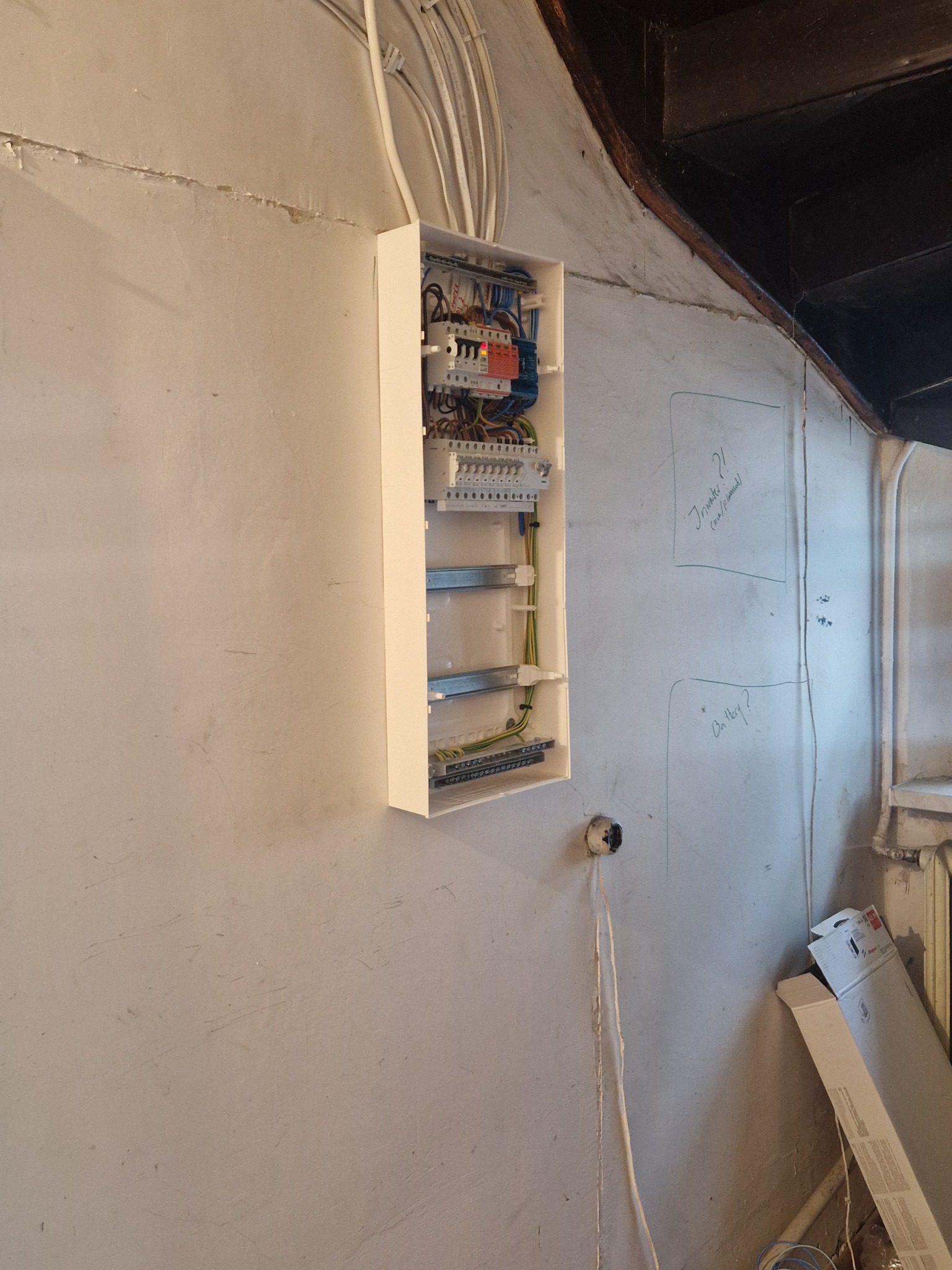 Otwarta skrzynka elektryczna z widocznymi przewodami i bezpiecznikami zamontowana na szarej, nierównej ścianie z notatkami i wystającymi kablami.