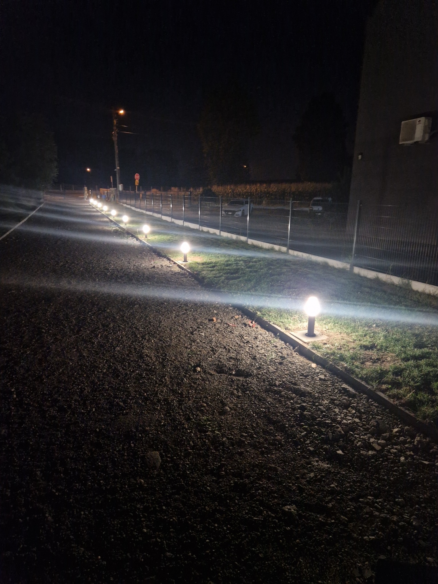 Rząd kulistych lamp oświetlających żwirową drogę wzdłuż ogrodzenia posesji w nocnym krajobrazie.
