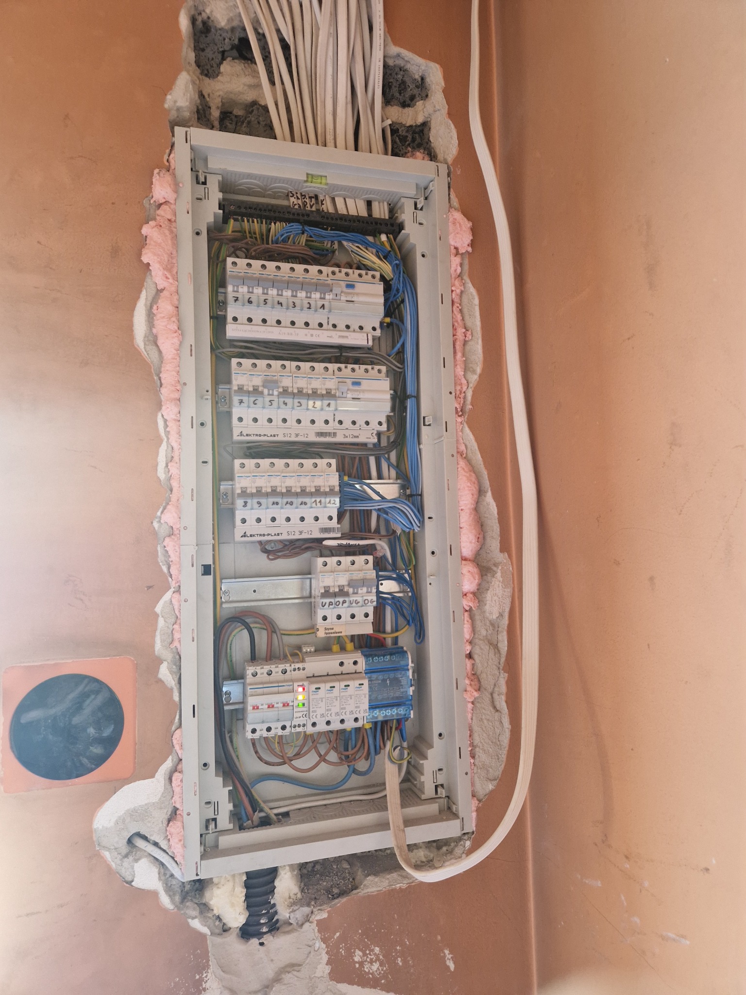 Rozdzielnica elektryczna w trakcie montażu, widoczne przewody, zabezpieczenia i różnicówka, ściana w kolorze łososiowym z różową pianką montażową wokół obudowy.