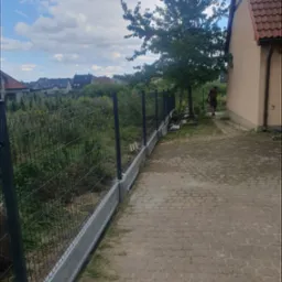 Nowo postawione ogrodzenie panelowe z podmurówką betonową na posesji, widok z boku, w tle domy jednorodzinne i pochmurne niebo.