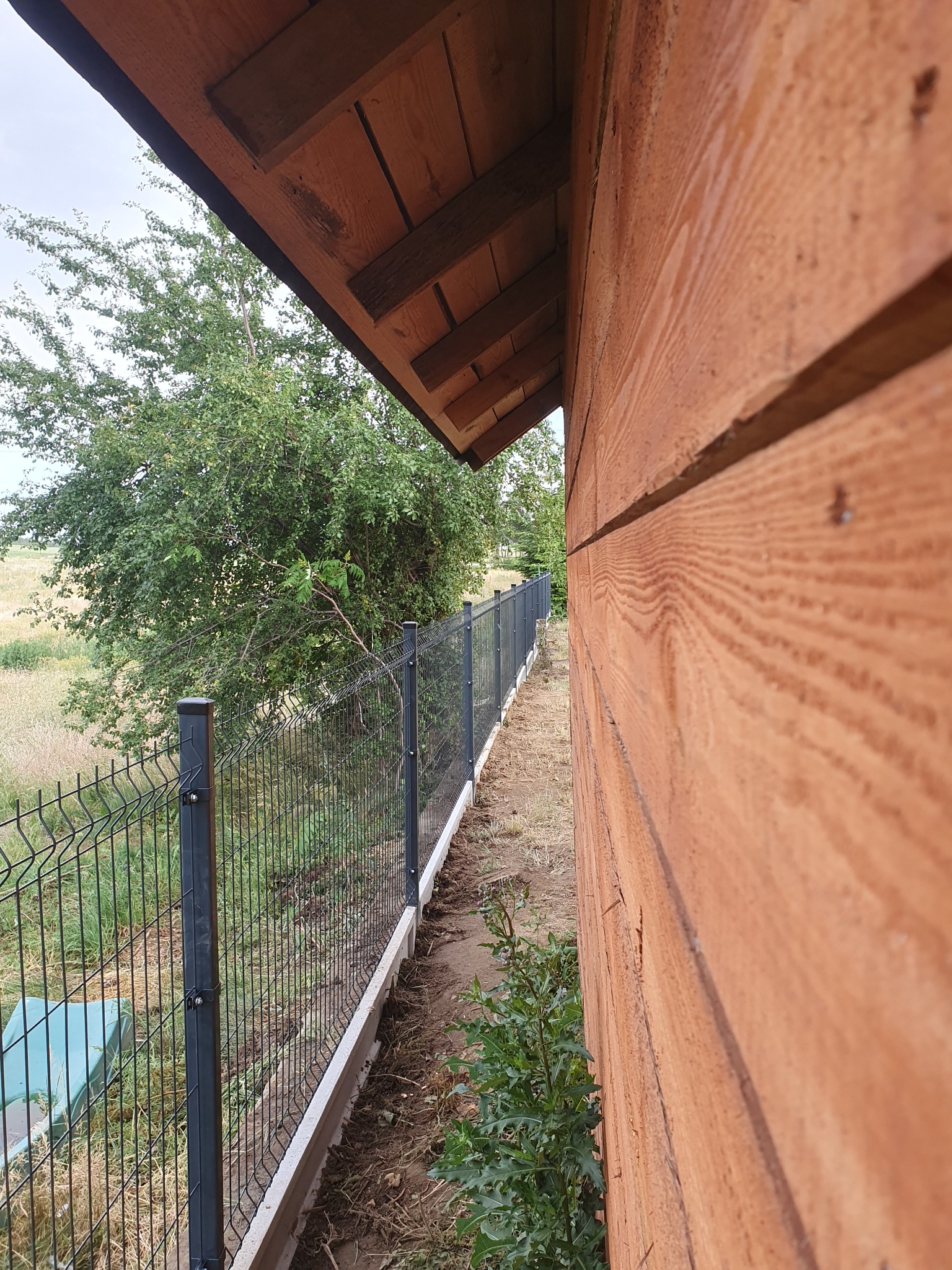 Nowoczesne, czarne ogrodzenie panelowe z podmurówką, widziane z perspektywy narożnika drewnianego budynku, z zielenią w tle.