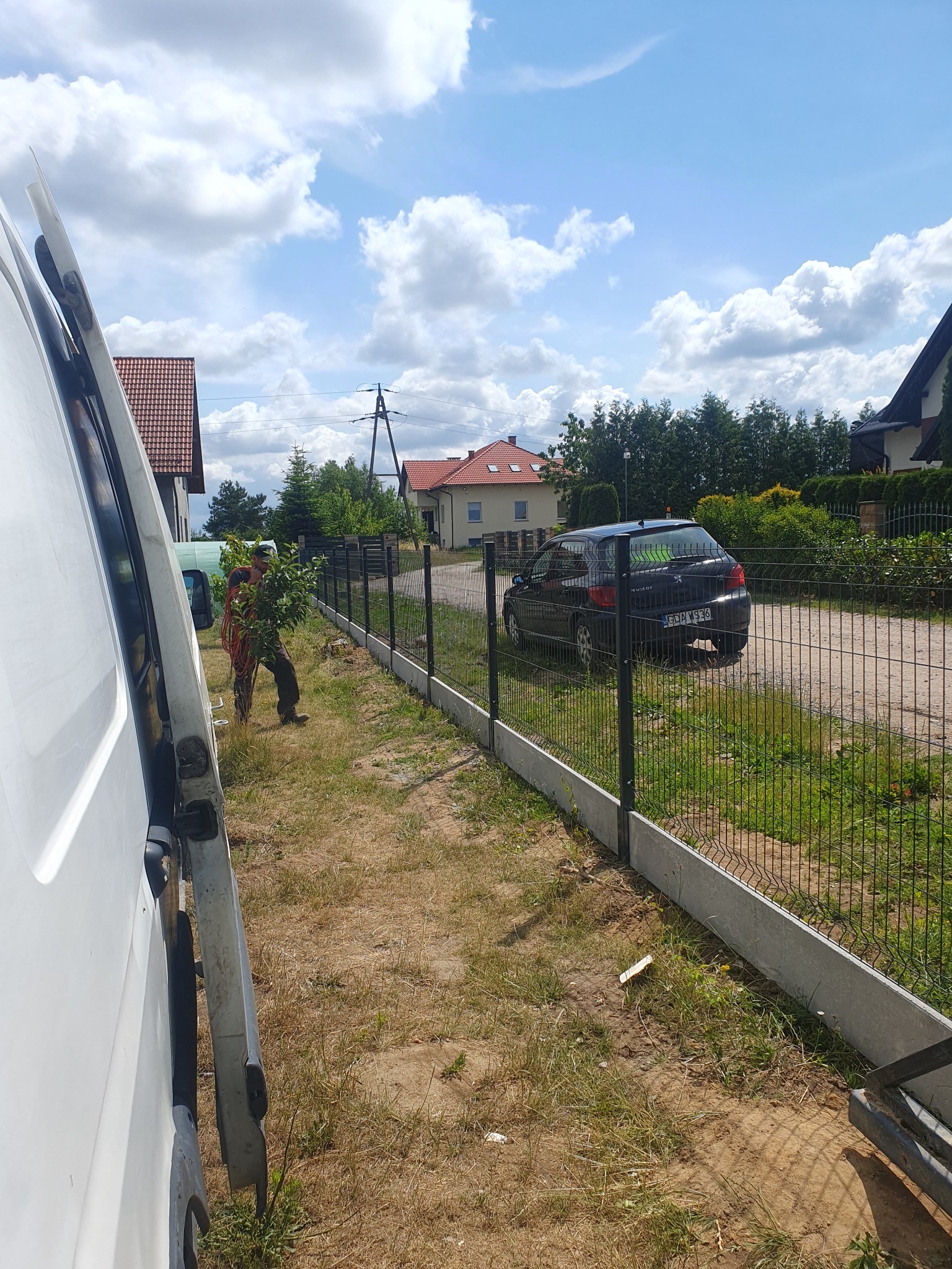 Pracownik z gałęziami w rękach stoi przy nowo postawionym, panelowym ogrodzeniu z podmurówką, obok zaparkowany samochód osobowy, widoczny samochód dostawczy z otwartymi drzwiami.