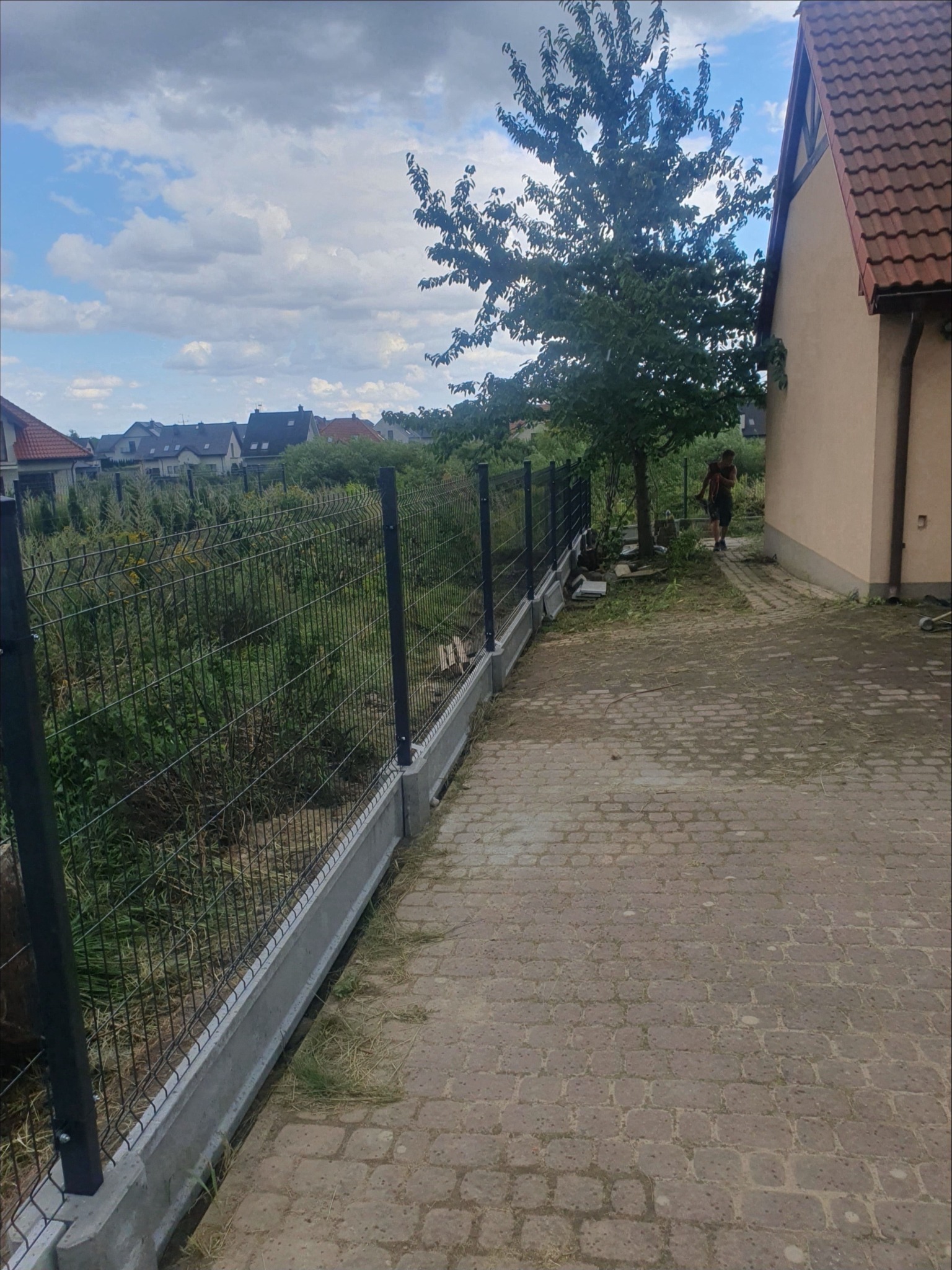 Nowo postawione ogrodzenie panelowe z podmurówką betonową na posesji, widok z boku, w tle domy jednorodzinne i pochmurne niebo.