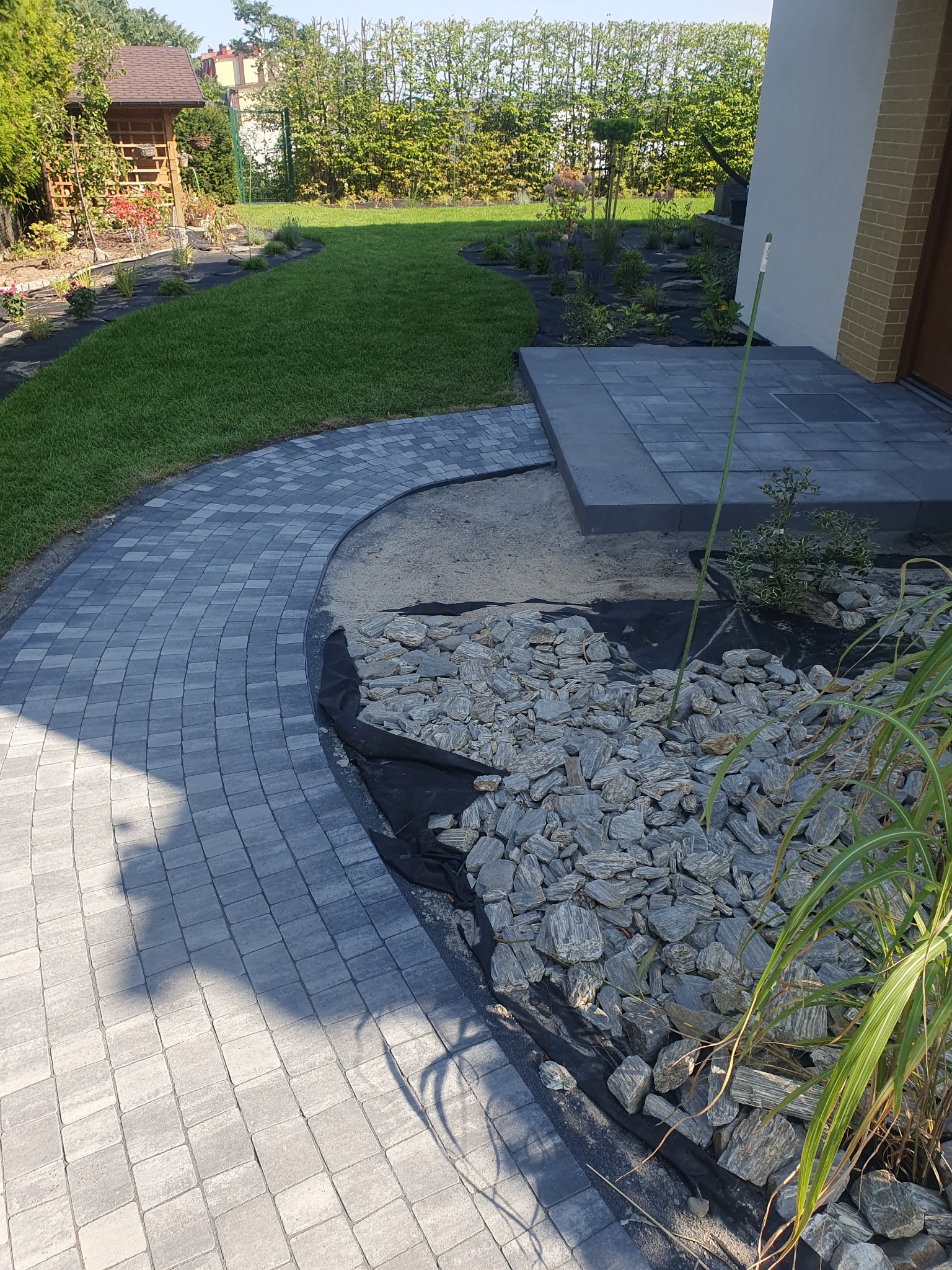 Ścieżka z kostki brukowej w kształcie łuku, obok rabata z kamieniami i czarną agrowłókniną, trawnik i roślinność ogrodowa w tle, widok z góry.