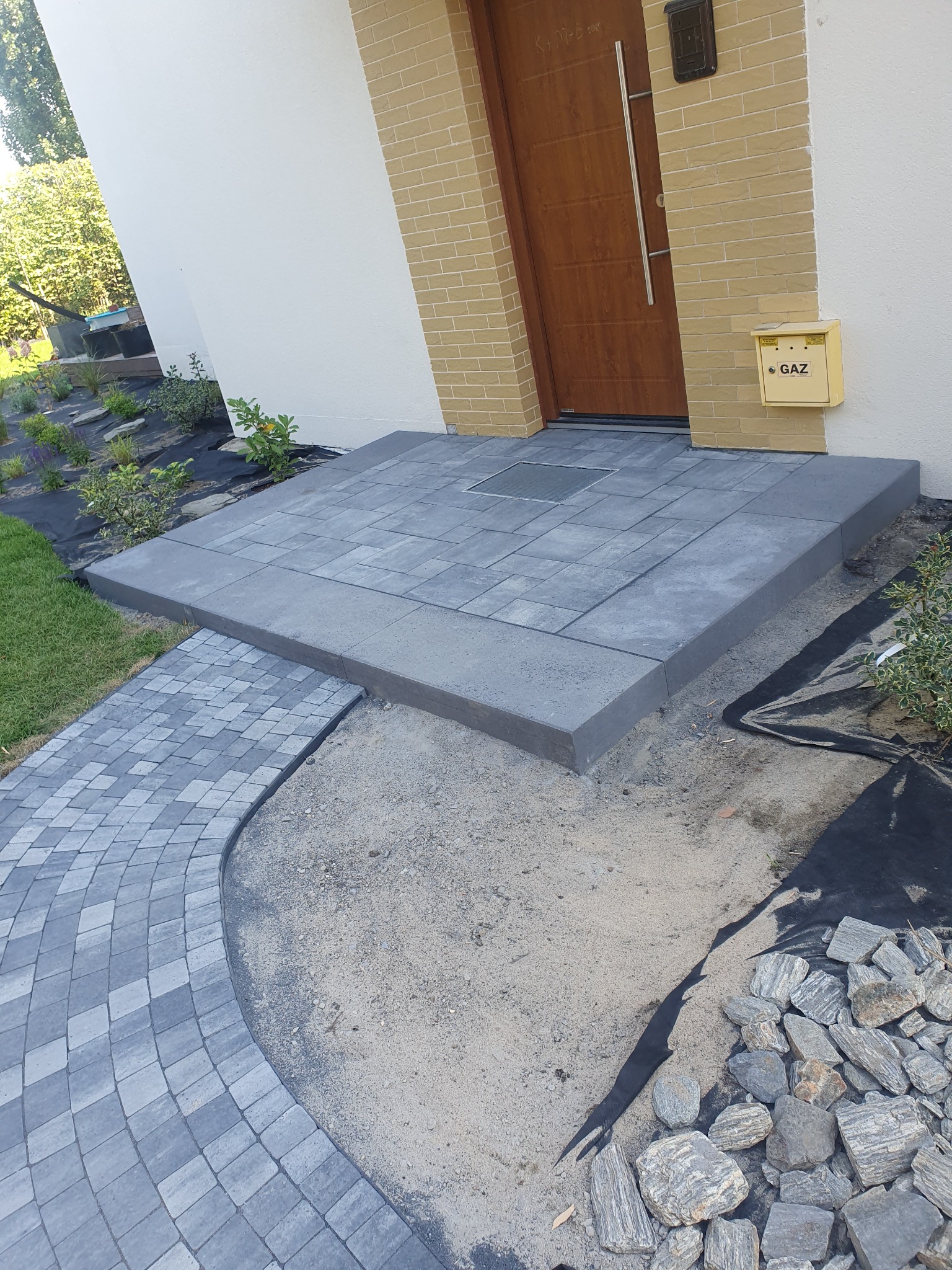 Ułożona kostka brukowa tworząca podest przed wejściem do domu, z widocznym fragmentem ścieżki z kostki, obok skrzynka gazowa i kamienie dekoracyjne.