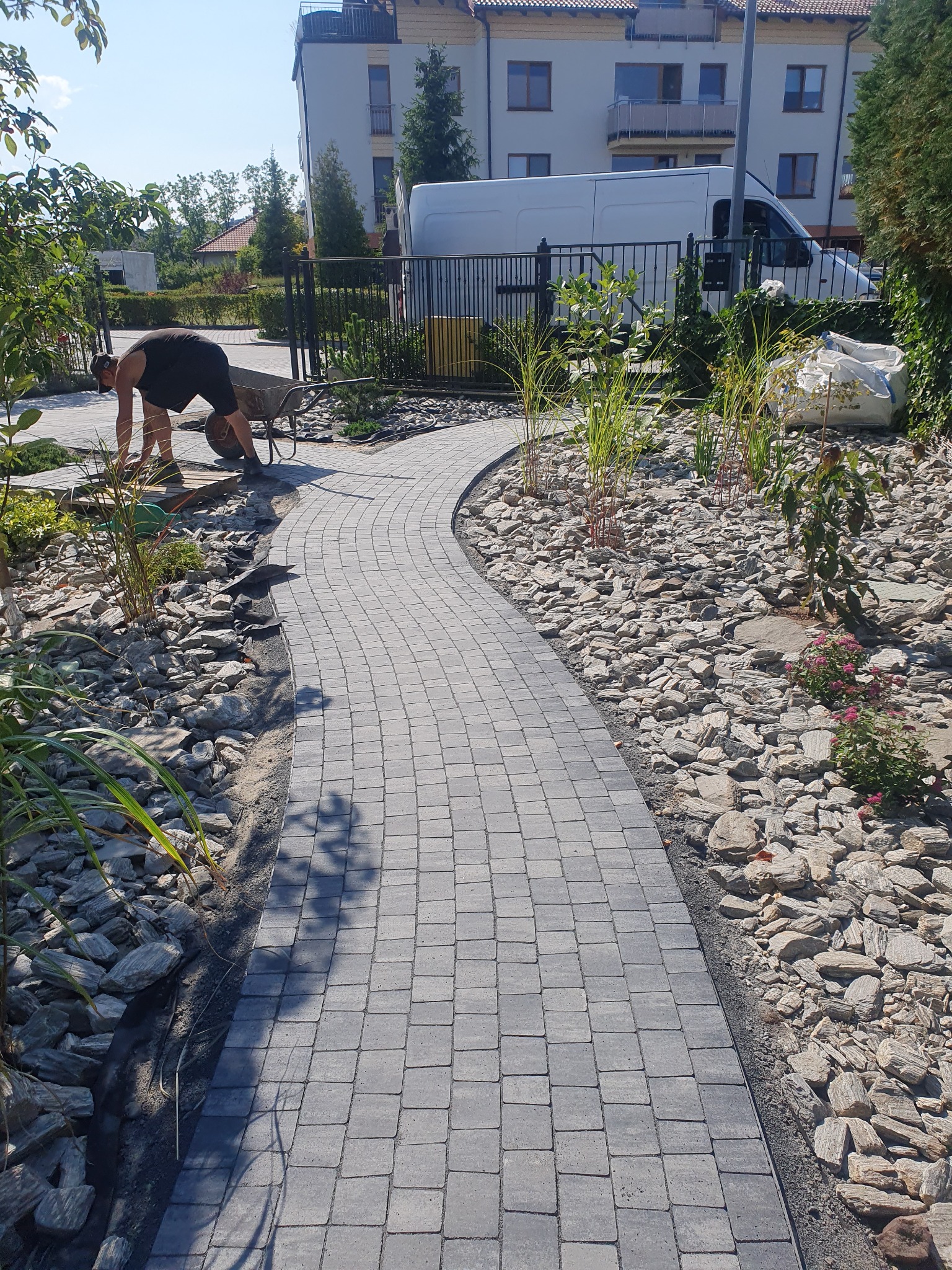 Układanie kostki brukowej na zakrzywionym chodniku w ogrodzie z elementami dekoracyjnymi z kamieni i roślin, pracownik z taczką w tle.