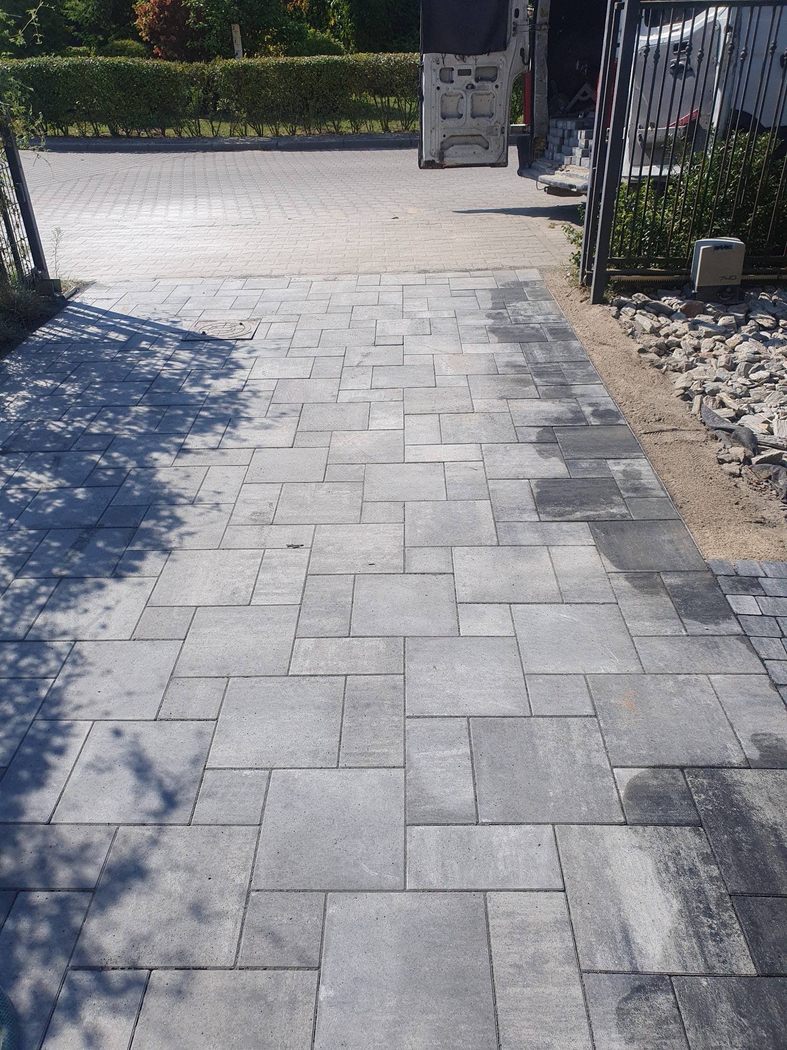 Świeżo ułożona, szara kostka brukowa na podjeździe, z widocznymi śladami wilgoci i cieniem drzewa, obok zaparkowany samochód dostawczy i brama wjazdowa.