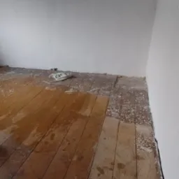 Drewniana podłoga po remoncie, pokryta plamami farby i resztkami gipsu, widoczna torba budowlana w rogu pomieszczenia.