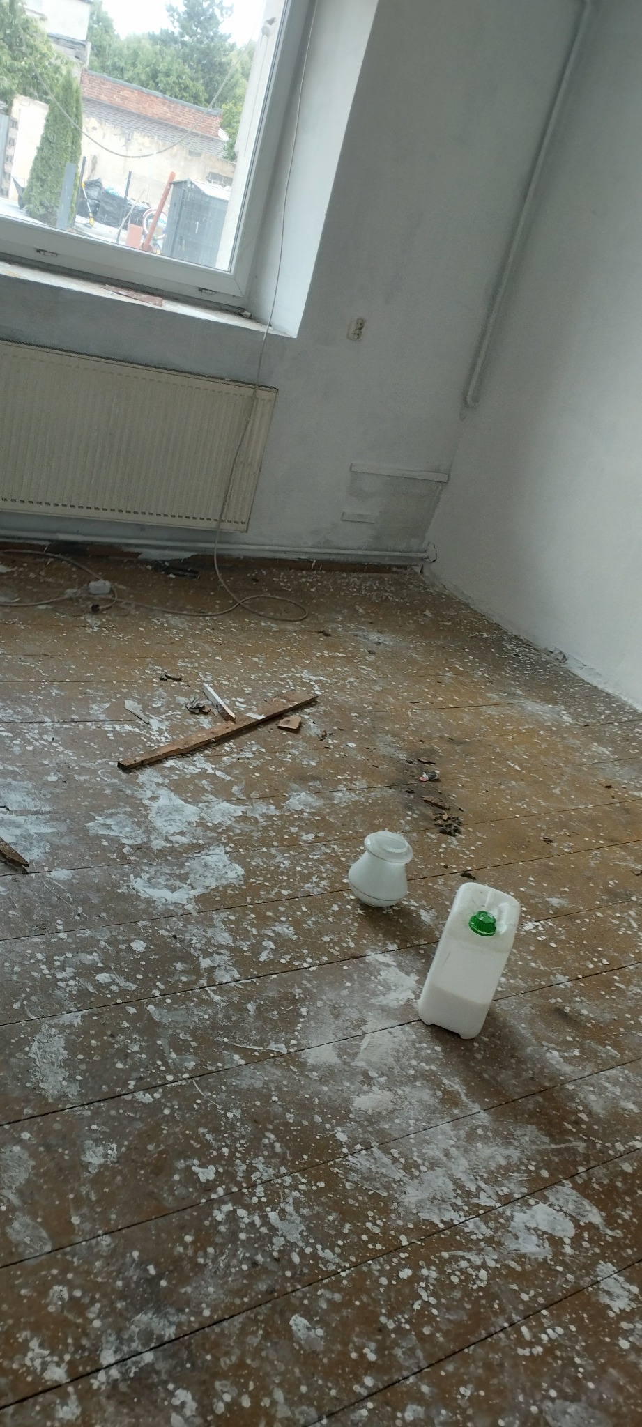 Podłoga z drewnianych desek pokryta plamami farby, widoczne resztki gruzu, butelka z płynem i szklany pojemnik, okno dachowe w tle.