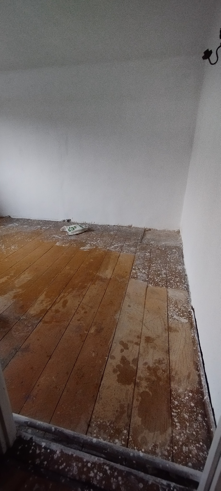 Drewniana podłoga po remoncie, pokryta plamami farby i resztkami gipsu, widoczna torba budowlana w rogu pomieszczenia.