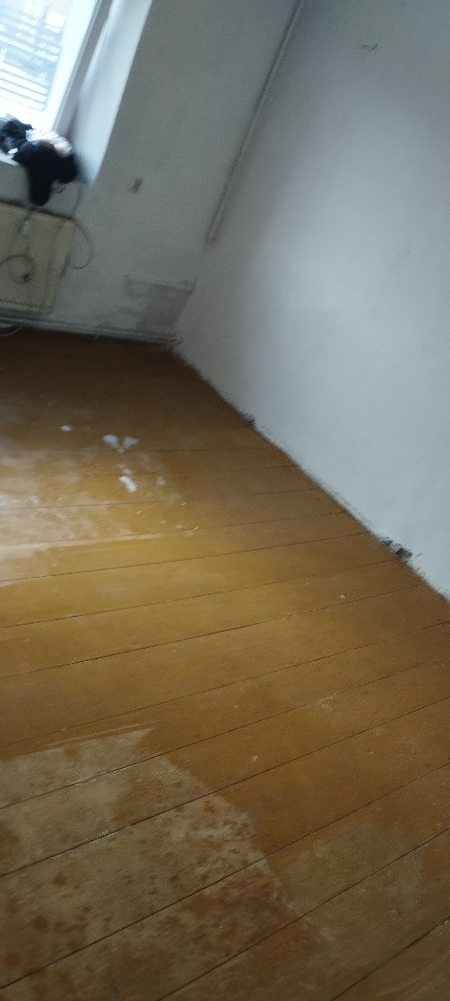 Pokój ze starą drewnianą podłogą, widoczne ślady po remoncie i niedokładnym sprzątaniu, białe ślady na podłodze, okno na poddaszu.