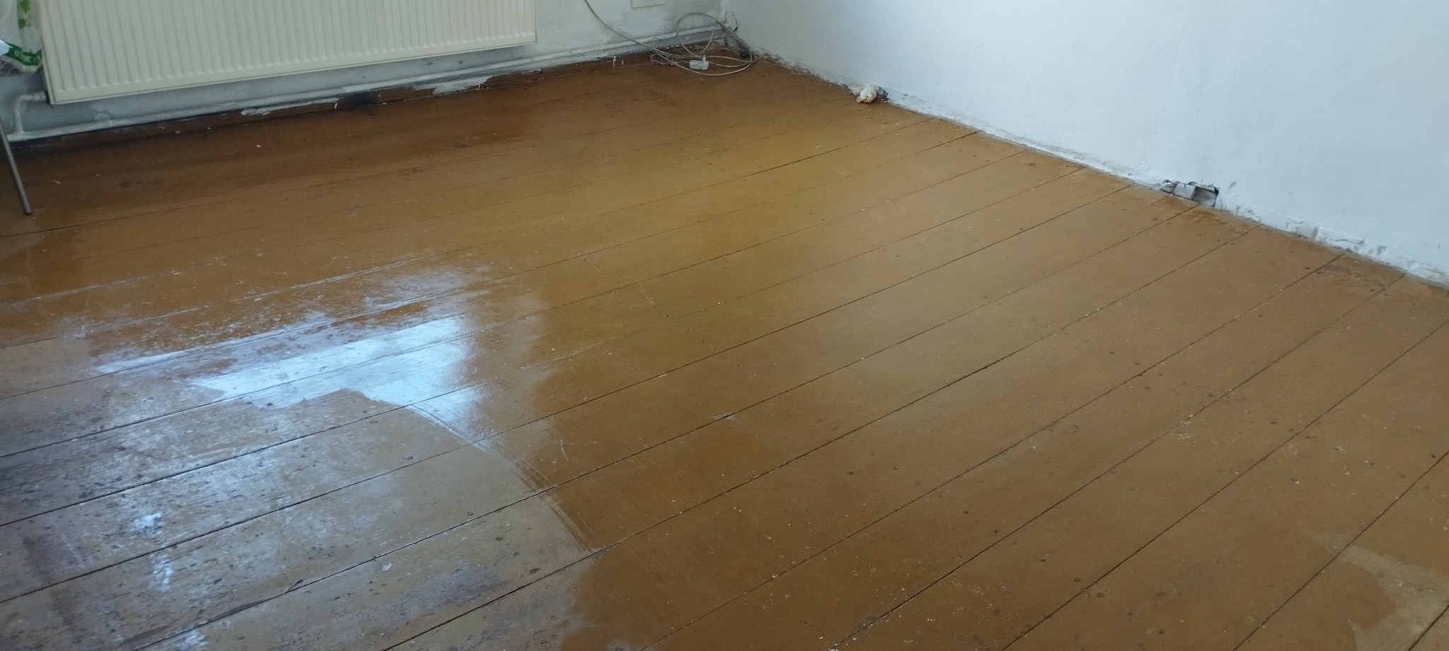 Świeżo umyta, mokra podłoga z desek w pokoju, widoczny grzejnik i kable przy ścianie.