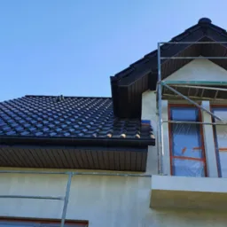 Dom jednorodzinny w trakcie prac elewacyjnych, z oknami zabezpieczonymi folią i ustawionym rusztowaniem; widoczna świeżo położona warstwa tynku w kolorze kremowym oraz czarna dachówka ceramiczna.