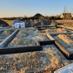 Wykopane i częściowo wymurowane fundamenty pod budynek jednorodzinny, widoczne bloczki Solbet, hałda piachu i ziemi, w tle zabudowa wiejska.