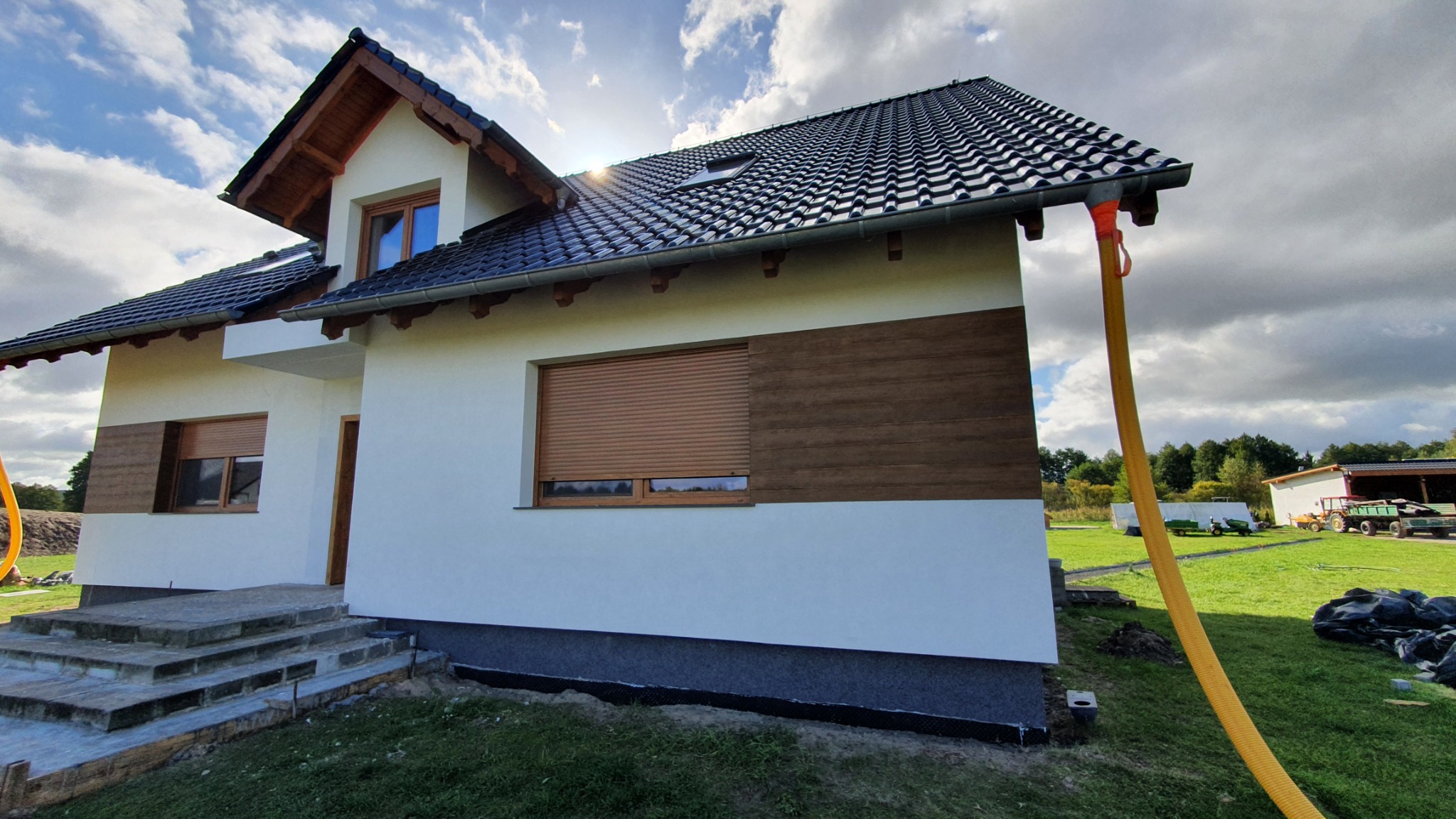 Nowo otynkowany dom jednorodzinny z drewnianymi akcentami elewacyjnymi i czarną dachówką, widoczny wąż do zaprawy na tle słonecznego nieba.