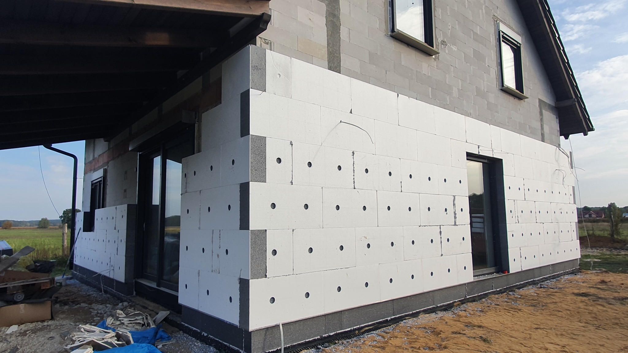 Dom w trakcie prac izolacyjnych: białe płyty styropianowe z otworami mocowane do szarego muru z bloczków. Widoczne okna, podbitka dachowa, fragment krajobrazu w tle.