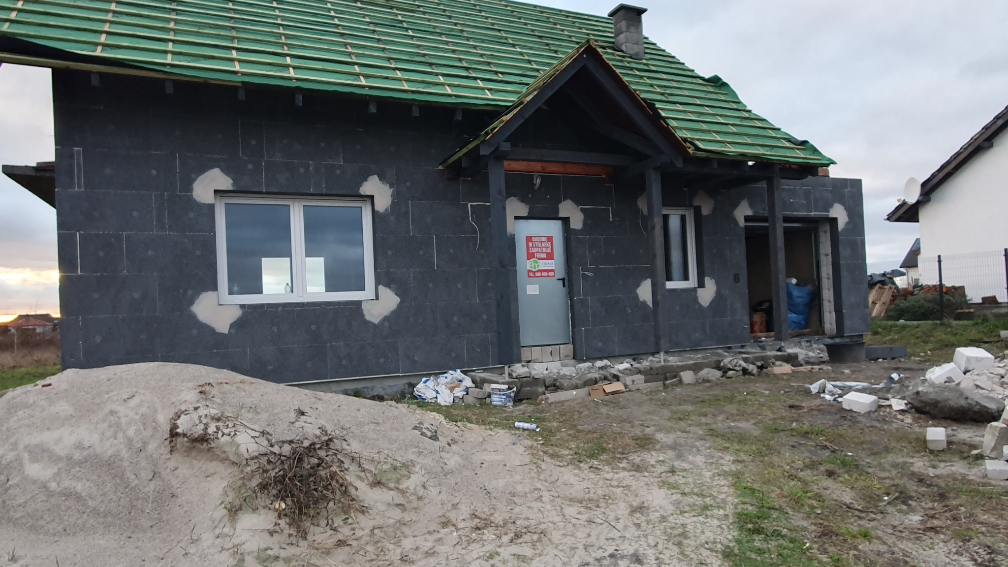 Dom jednorodzinny w trakcie prac: ściany zewnętrzne pokryte szarą izolacją, częściowo zaszpachlowane, zielony dach z widoczną konstrukcją, na elewacji naklejona reklama firmy, na pierwszym planie...