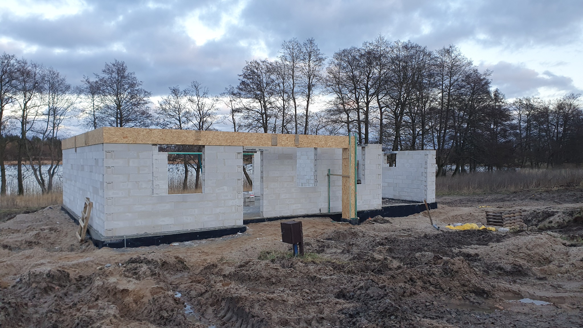 Budowa domu jednorodzinnego w stanie surowym otwartym z bloczków betonowych, widoczne fundamenty z izolacją, drewniana konstrukcja wieńca, tło z jeziorem i drzewami.