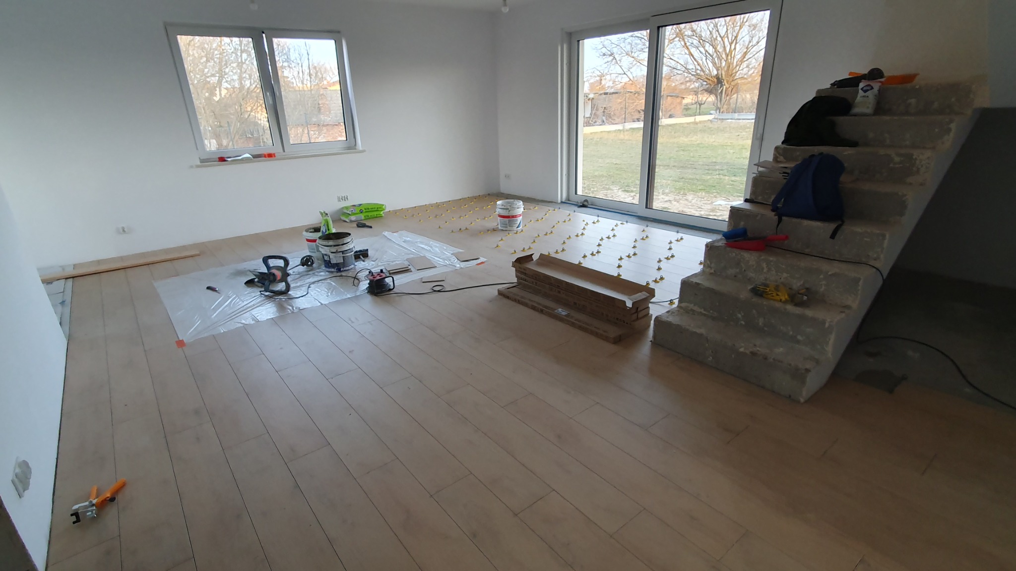 Układanie podłogi z paneli drewnopodobnych w jasnym, nowym wnętrzu; widoczne narzędzia, paczka paneli, poziomica, wiaderka z klejem i krzyżyki dystansowe.