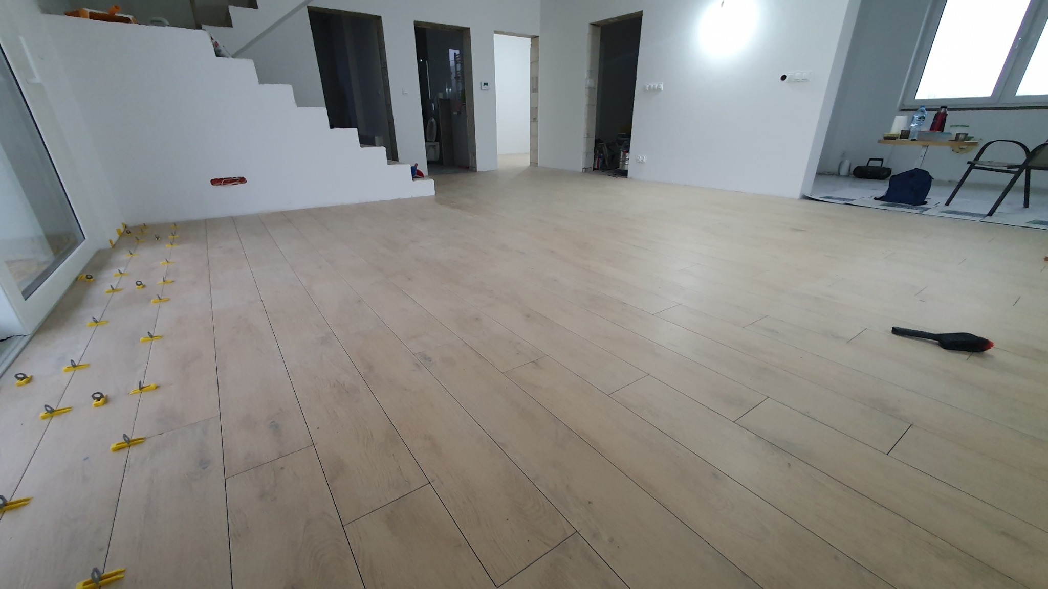 Układanie podłogi z paneli imitujących drewno, widoczne żółte klipsy poziomujące i szczotka do zamiatania, białe ściany i schody w tle.