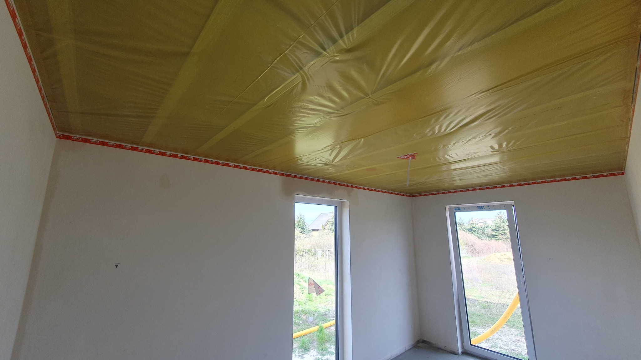 Wnętrze w Skwierzynie z folią paroizolacyjną na suficie, przygotowane do dalszych prac wykończeniowych. Widoczne surowe ściany i okna z widokiem na zewnątrz.