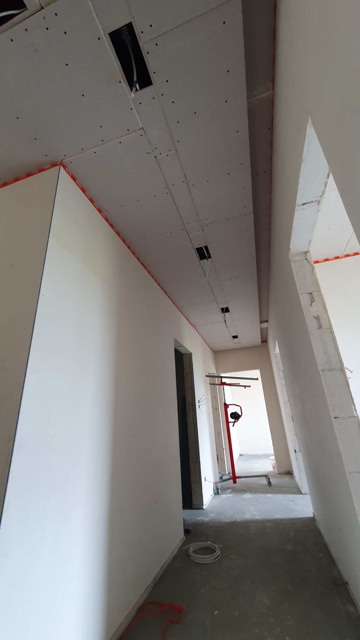 Wnętrze w trakcie prac wykończeniowych: widoczne płyty gipsowo-kartonowe na suficie z otworami na instalacje, ściany przygotowane do malowania, czerwona poziomica laserowa na statywie w tle,...