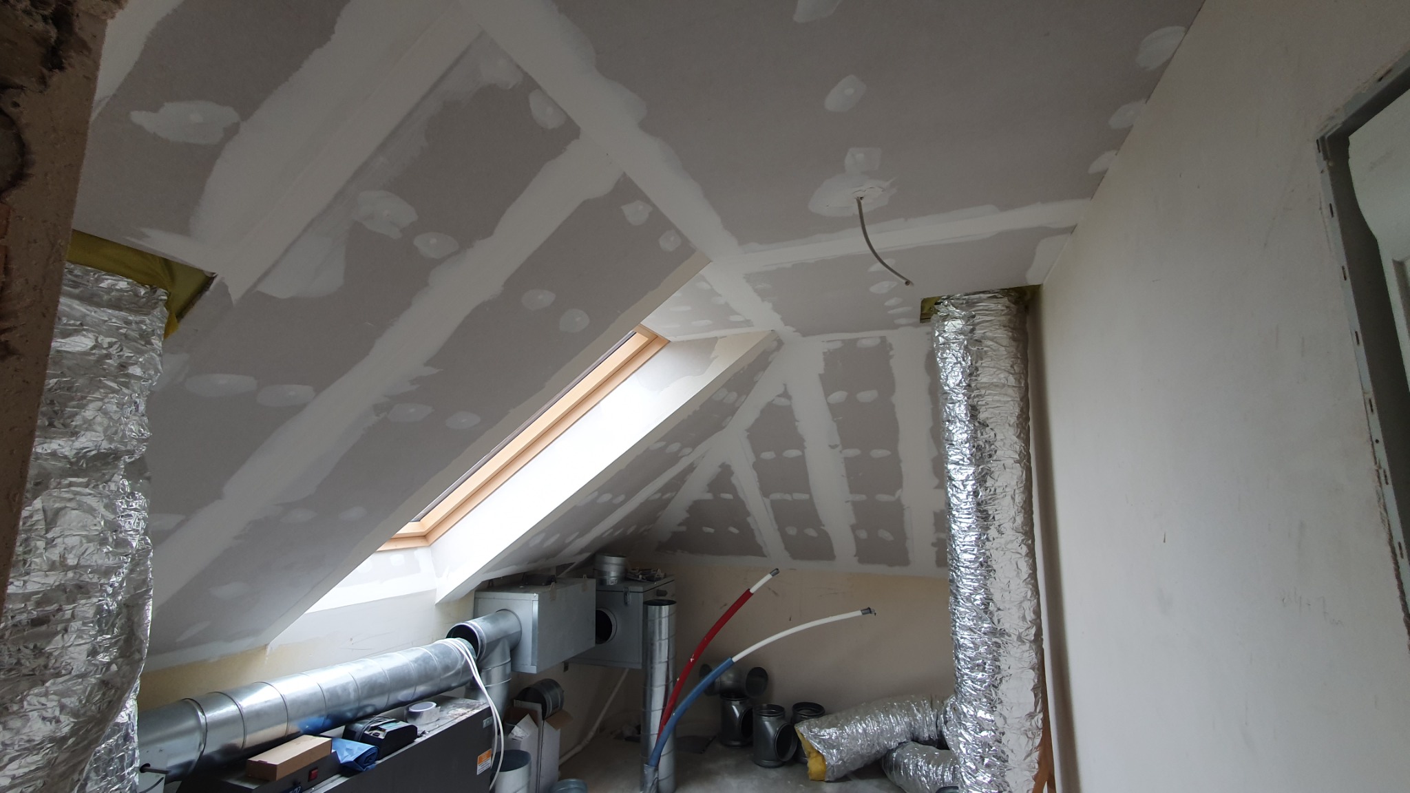 Poddasze w trakcie wykończenia: płyty gipsowo-kartonowe na skosach dachu z widocznym oknem dachowym, łączenia płyt zaszpachlowane, instalacja wentylacyjna z rurami aluminiowymi, widoczne elementy...