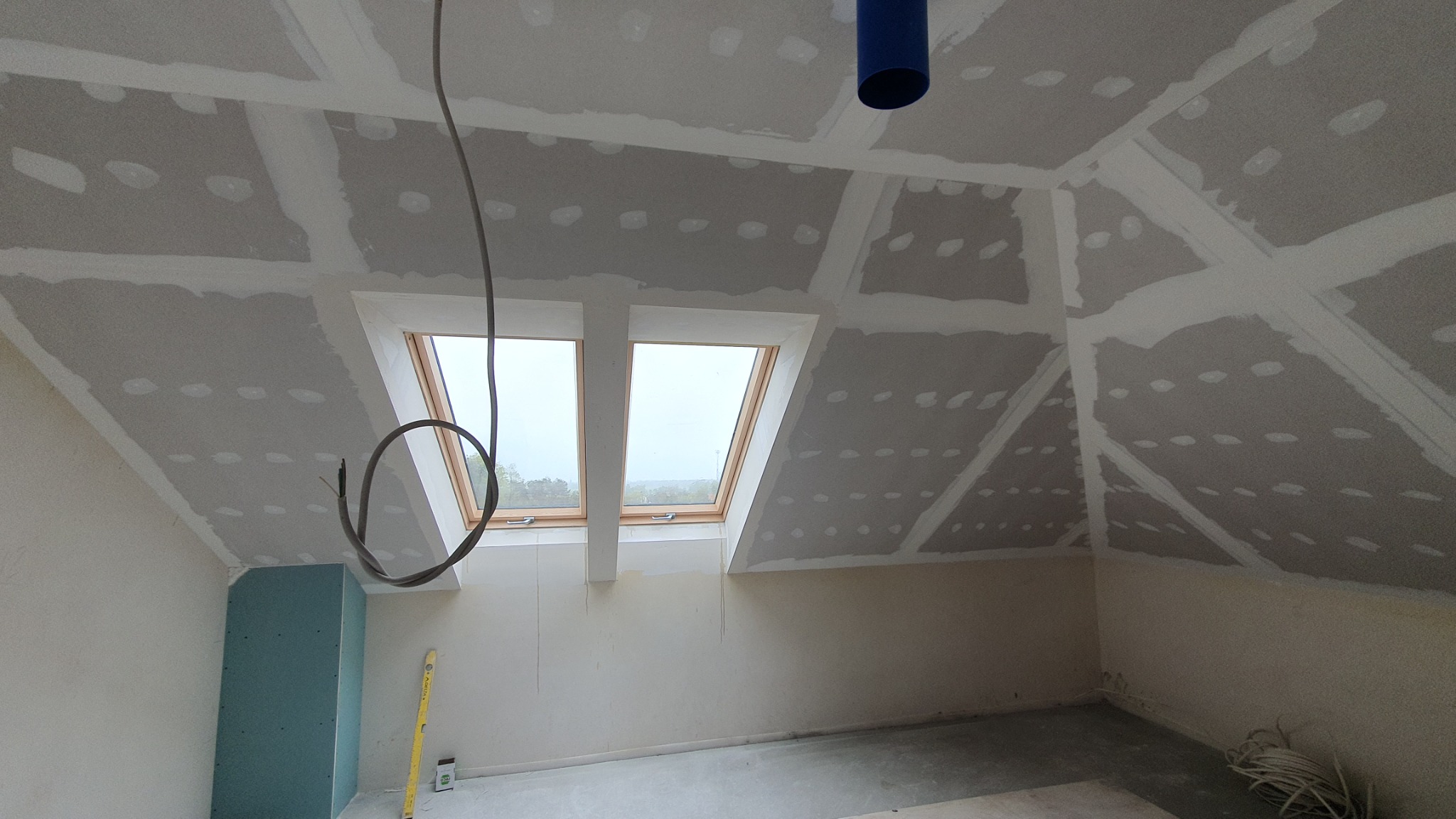 Poddasze z oknami dachowymi w trakcie prac wykończeniowych: płyty gipsowo-kartonowe na skosach i suficie, szpachlowane łączenia, wystające przewody elektryczne, poziomica oparta o ścianę