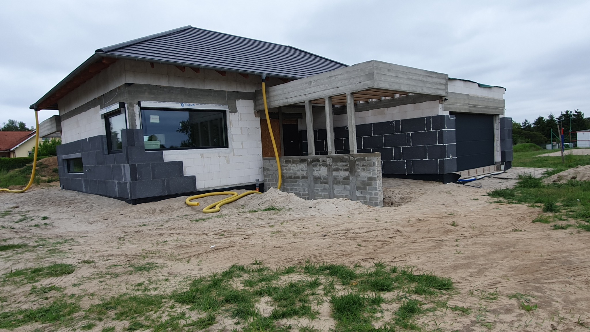 Surowy stan domu jednorodzinnego z szarej cegły z częściowo wykonaną izolacją fundamentów z czarnego styropianu, widoczne szare bloczki fundamentowe i żółty wąż doprowadzający wodę.